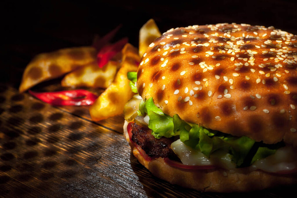 Burger with french fries on a dark wooden board. Cheeseburger with cutlet and herbs on black background. Food with contrasting dramatic light. Potatoes with red peppers closeup. - Photo, Image