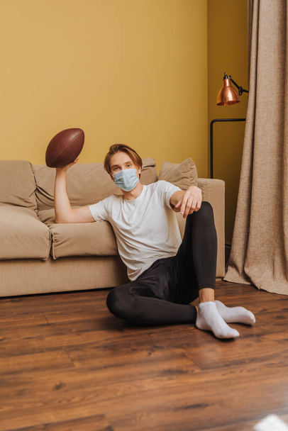 young man in medical mask holding american football while sitting on floor in living room  - 写真・画像