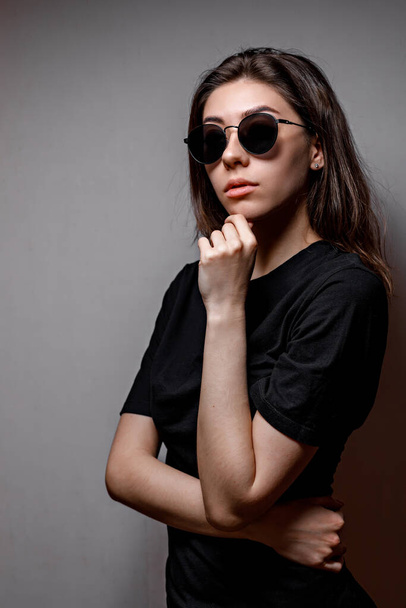 hermosa mujer morena joven en gafas de sol negras y una camiseta negra sobre un fondo gris
 - Foto, Imagen