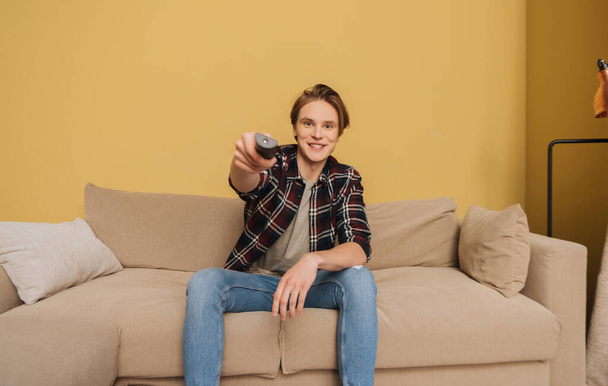 Junger lächelnder Mann hält Fernbedienung in der Hand und sitzt auf Sofa, Ende der Quarantäne  - Foto, Bild