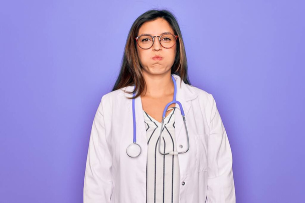 Médecin professionnel femme portant stéthoscope et manteau médical sur fond violet joues gonflantes avec visage drôle. Bouche gonflée avec de l'air, expression folle
. - Photo, image