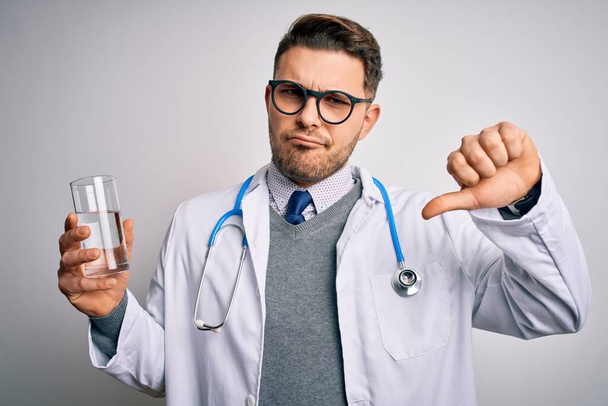 Νεαρός γιατρός που φοράει ιατρική ποδιά κρατώντας ένα ποτήρι φρέσκο νερό σε απομονωμένο φόντο με θυμωμένο πρόσωπο, αρνητική ένδειξη που δείχνει αντιπάθεια με τους αντίχειρες κάτω, έννοια απόρριψης - Φωτογραφία, εικόνα