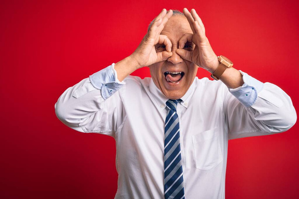 Senior knappe zakenman draagt elegante stropdas staande over geïsoleerde rode achtergrond doen ok gebaar als verrekijker steken tong uit, ogen kijken door vingers. Gekke uitdrukking. - Foto, afbeelding