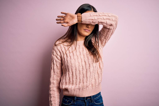 Junge schöne brünette Frau in lässigem Pullover über isoliertem rosa Hintergrund, der die Augen mit dem Arm bedeckt, sieht ernst und traurig aus. Sichtloses, verstecktes und verwerfliches Konzept - Foto, Bild