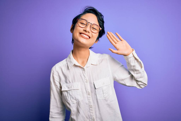 Jeune belle fille asiatique portant une chemise décontractée et des lunettes debout sur fond violet Renonçant à dire bonjour heureux et souriant, geste de bienvenue amical
 - Photo, image