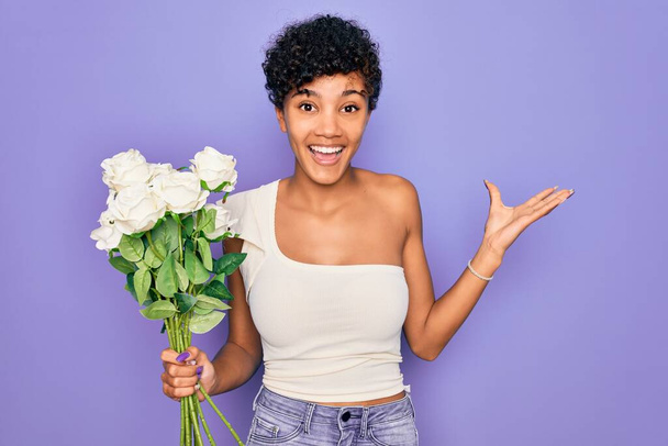Mooie Afrikaanse amerikaanse afro vrouw met boeket van witte bloemen over paarse achtergrond zeer gelukkig en opgewonden, winnaar uitdrukking vieren overwinning schreeuwen met grote glimlach en opgeheven handen - Foto, afbeelding