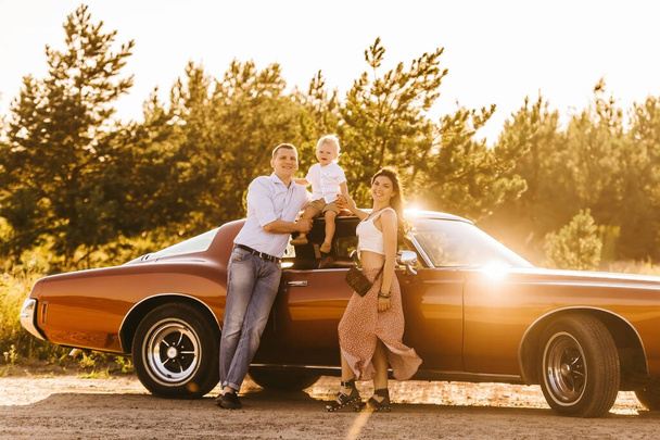 Buick Riviera in retro style. Unique car. Cute blond boy sitting on the roof of the car. Parents hug their son at sunset - Foto, afbeelding