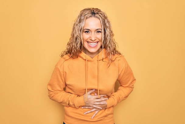 Young beautiful blonde sporty woman wearing casual sweatshirt over yellow background smiling and laughing hard out loud because funny crazy joke with hands on body. - Photo, Image