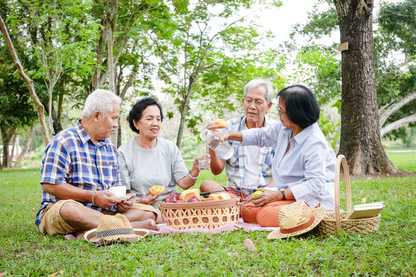 A group of Asian elders are sitting, relaxing and preparing snacks to eat in the shady garden. Watch them happy. Concepts of the elderly community. Picnic at the park - Photo, Image