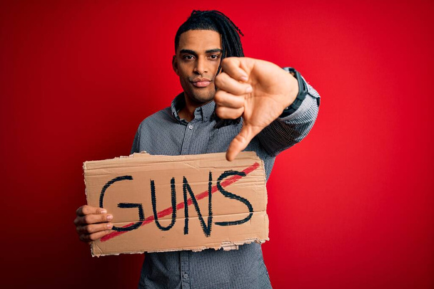 Joven afroamericano con rastas sosteniendo pancarta con armas prohibidas protesta con la cara enojada, signo negativo que muestra aversión con los pulgares hacia abajo, concepto de rechazo
 - Foto, Imagen
