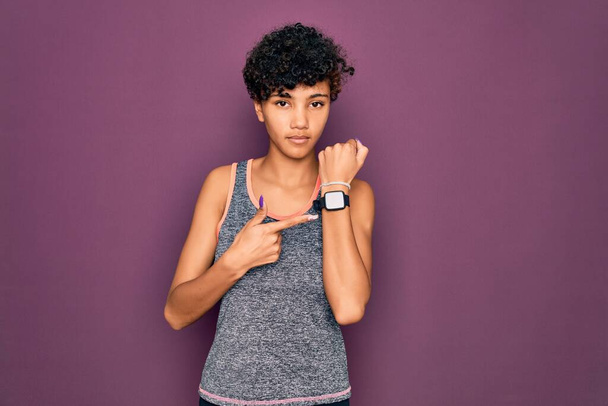 Young beautiful african american afro sportswoman doing exercise wearing sportswear In hurry pointing to watch time, impatience, looking at the camera with relaxed expression - Photo, Image