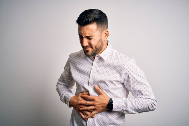 Young handsome man wearing elegant shirt standing over isolated white background with hand on stomach because indigestion, painful illness feeling unwell. Ache concept. - Photo, Image