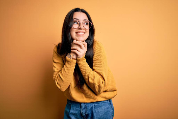 Young brunette woman wearing glasses and casual sweater over yellow isolated background laughing nervous and excited with hands on chin looking to the side - Foto, Bild