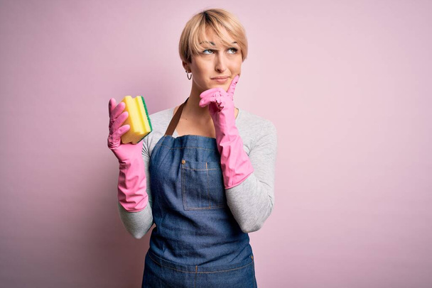 Young blonde cleaner woman with short hair wearing apron and gloves holding scrub scourer serious face thinking about question, very confused idea - Фото, изображение