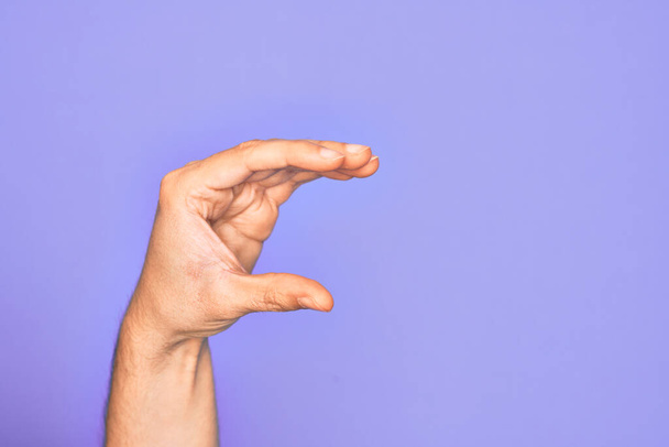 Ruka kavkazského mladíka ukazující prsty na izolovaném purpurovém pozadí vybírající a užívající neviditelnou věc, držící objekt s prsty ukazujícími prostor - Fotografie, Obrázek