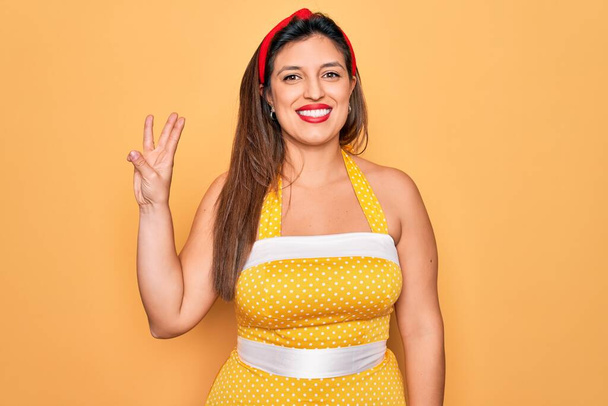 Junge hispanische Pin-up-Frau trägt Mode sexy 50er Jahre Stil auf gelbem Hintergrund zeigt und zeigt mit Fingern Nummer drei, während sie selbstbewusst und glücklich lächelt. - Foto, Bild