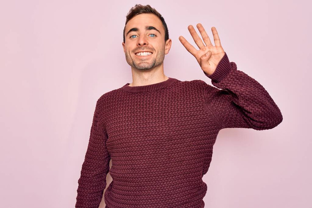 Young handsome man with blue eyes wearing casual sweater standing over pink background showing and pointing up with fingers number four while smiling confident and happy. - Фото, изображение
