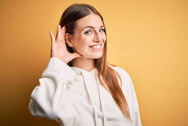 Jeune belle femme sportive rousse portant un sweat-shirt sur fond jaune isolé souriant avec main sur oreille en écoutant une rumeur ou des commérages. Concept de surdité
. - Photo, image