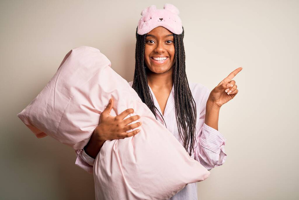 Young african american woman wearing pajama and eye mask hugging bed pillow very happy pointing with hand and finger to the side - Фото, изображение