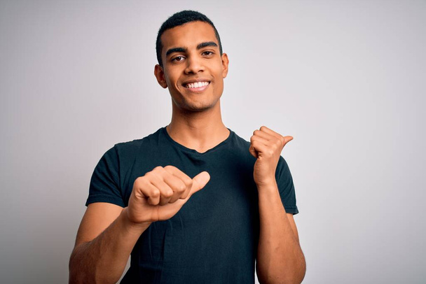Jeune bel homme afro-américain portant un t-shirt décontracté debout sur fond blanc Pointant vers le dos derrière avec la main et les pouces levés, souriant confiant
 - Photo, image