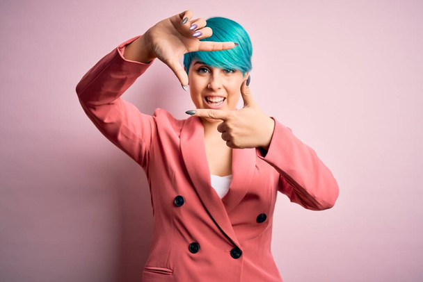 Молода красива бізнес-леді з синім модним волоссям в куртці на рожевому фоні, посміхаючись роблячи рамку руками і пальцями з щасливим обличчям. Концепція творчості та фотографії
. - Фото, зображення