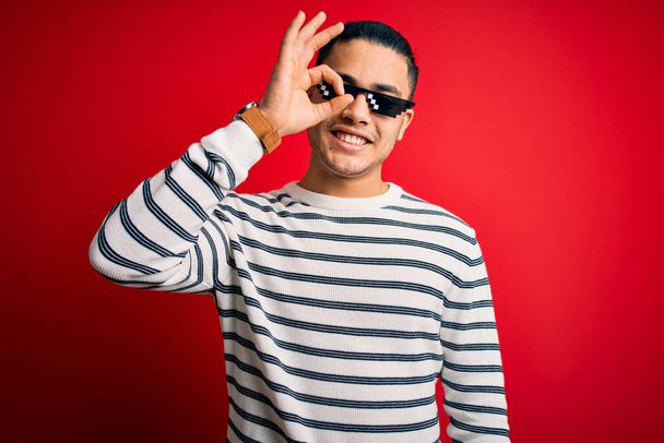 Jeune homme brésilien portant des lunettes de soleil de vie de voyou drôle sur fond rouge isolé faisant un geste correct avec la main souriant, oeil regardant à travers les doigts avec un visage heureux
. - Photo, image