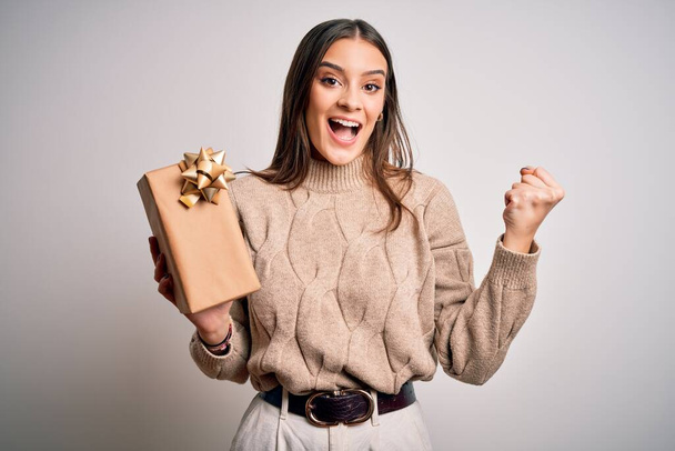 Young beautiful brunette woman holding birthday gift over isolated white background screaming proud and celebrating victory and success very excited, cheering emotion - Valokuva, kuva