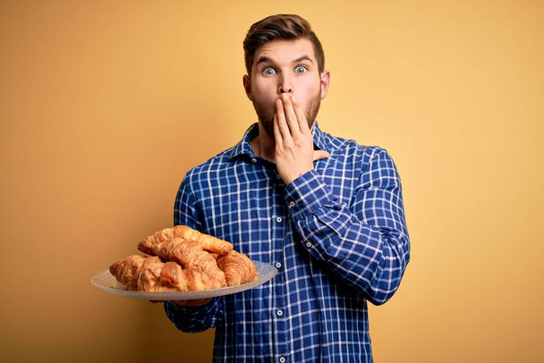 Young blond man with beard and blue eyes holding plate with french croissants to breakfast cover mouth with hand shocked with shame for mistake, expression of fear, scared in silence, secret concept - Photo, Image