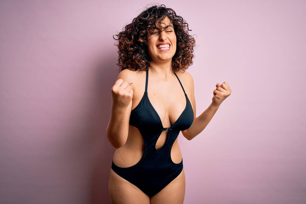 Young beautiful arab woman on vacation wearing swimsuit and sunglasses over pink background excited for success with arms raised and eyes closed celebrating victory smiling. Winner concept. - Fotografie, Obrázek