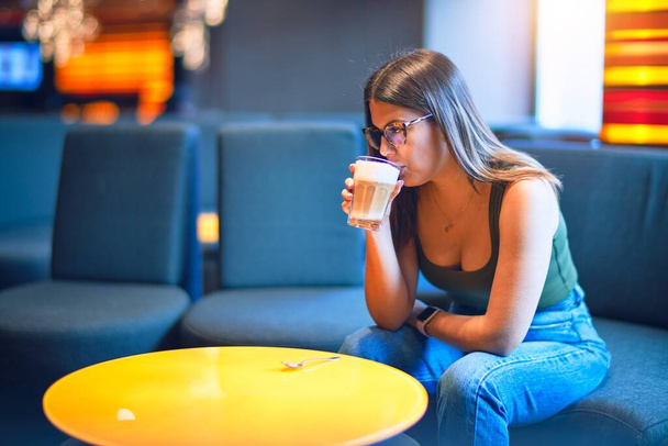 Young beautiful woman drinking glass of coffee sitting at restaurant - 写真・画像