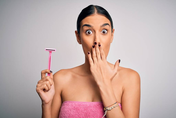 Young beautiful brunette woman wearing towel using razor for depilation cover mouth with hand shocked with shame for mistake, expression of fear, scared in silence, secret concept - Photo, Image
