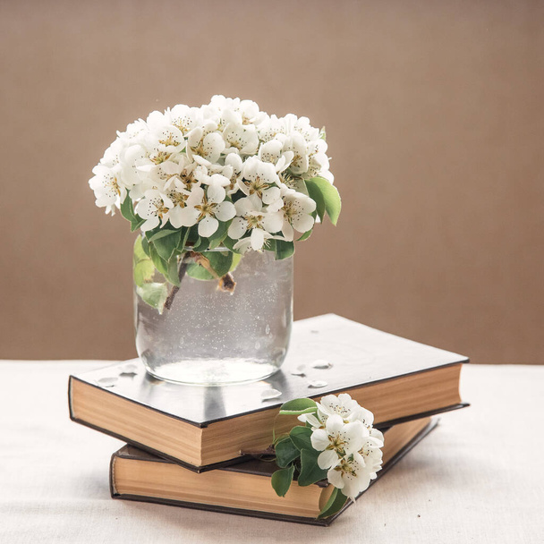 Blossom pear bouquet in a glass jar is on a brown old books on beige linen tablecloth. Spring rustic composition with white flowers. Romantic seasonal spring decoration ideas for your interior. - Foto, immagini