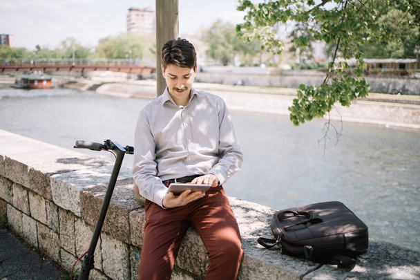 Freelancer man using tablet while sitting on stone fence near river - 写真・画像