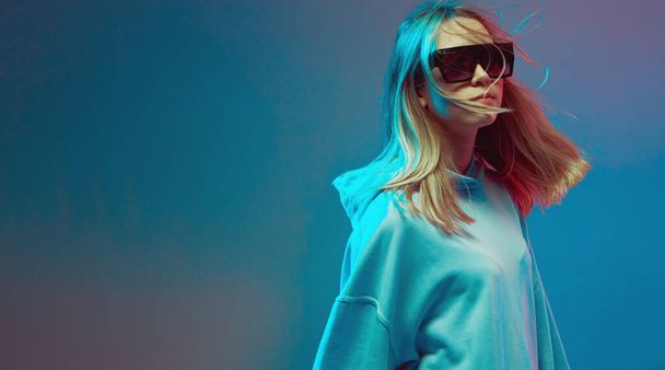 Portrait of a stylish young girl, cool posing in a hoodie, sunglasses and with developing hair, on a neon background. - Photo, image