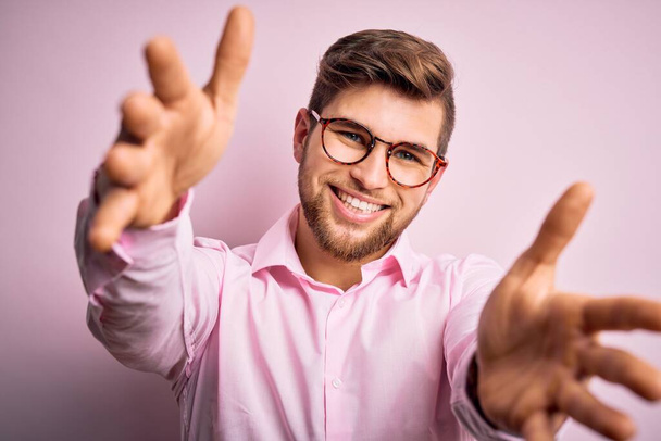 Young handsome blond man with beard and blue eyes wearing pink shirt and glasses looking at the camera smiling with open arms for hug. Cheerful expression embracing happiness. - Photo, Image