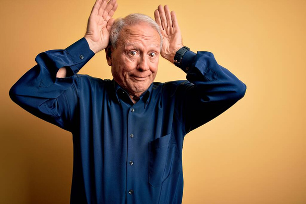 Grey haired senior man wearing casual blue shirt standing over yellow background Doing bunny ears gesture with hands palms looking cynical and skeptical. Easter rabbit concept. - Photo, Image
