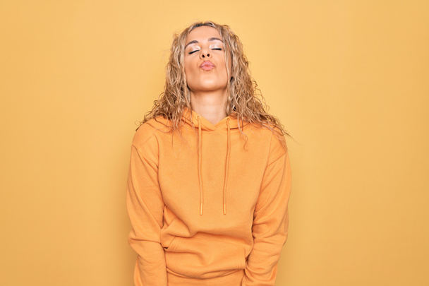Junge schöne, blonde, sportliche Frau in lässigem Sweatshirt vor gelbem Hintergrund, die in die Kamera blickt und einen Kuss in die Luft bläst, ist schön und sexy. Liebesbekundung. - Foto, Bild