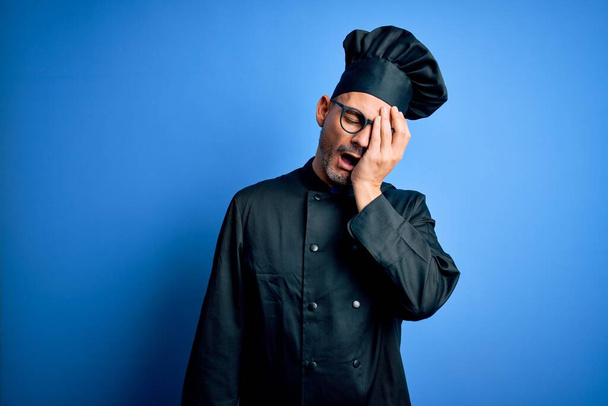 Mladý pohledný kuchař v kuchařské uniformě a klobouku na osamělém modrém pozadí Zívající unaveně pokrývající polotvář, oko a ústa rukou. Tvář bolí bolestí. - Fotografie, Obrázek