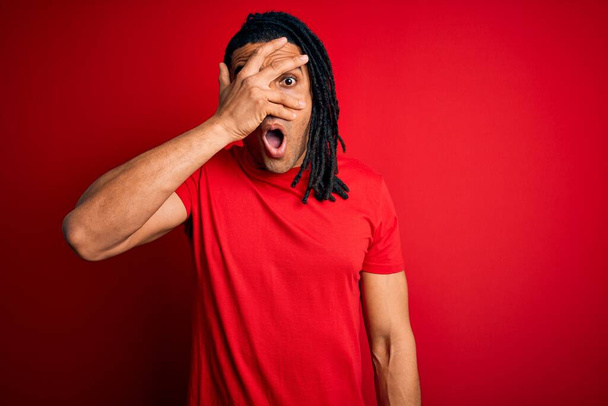 Jonge knappe Afrikaans-Amerikaanse afro man met dreadlocks in rood casual t-shirt gluren in shock bedekken gezicht en ogen met de hand, kijkend door vingers met beschaamde uitdrukking. - Foto, afbeelding