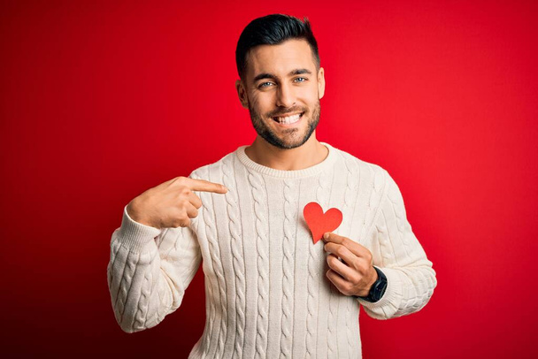 Jeune homme beau tenant coeur en forme de rouge comme symbole romantique et de santé avec visage surprise pointant du doigt vers lui-même
 - Photo, image