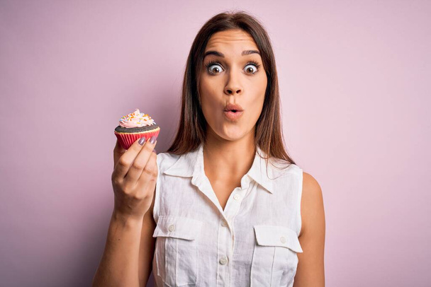 Jovem bela mulher morena comendo cupcake de chocolate sobre fundo rosa isolado assustado em choque com um rosto surpresa, com medo e animado com expressão de medo
 - Foto, Imagem