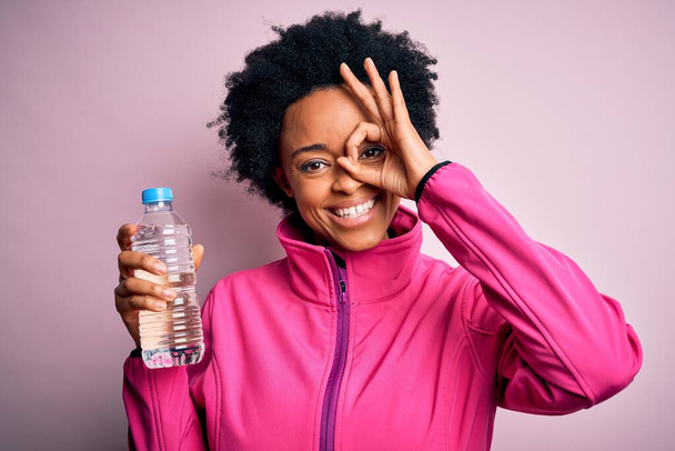 Молода афроамериканська спортсменка з кучерявим волоссям, що п'є пляшку води з щасливим обличчям, посміхається, роблячи знак рукою на око, дивлячись крізь пальці. - Фото, зображення