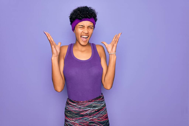 Beautiful african american afro woman wearing casual sportswear over purple background celebrating mad and crazy for success with arms raised and closed eyes screaming excited. Winner concept - Photo, Image