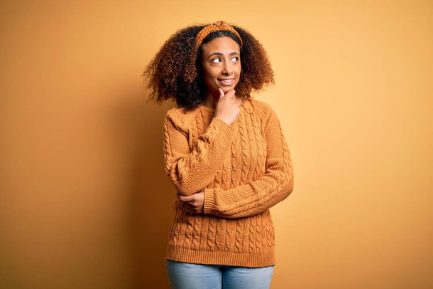 Nuori afrikkalainen amerikkalainen nainen, jolla on afro-hiukset yllään rento villapaita keltaisella taustalla, käsi leualla ajatellen kysymystä, mietteliäs ilme. Hymyillen ajattelevaisilla kasvoilla. Epävarma käsite
. - Valokuva, kuva