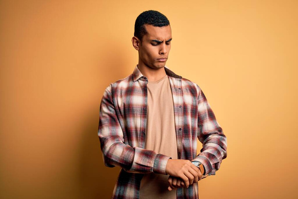 Mladý pohledný africký Američan muž na sobě ležérní košili stojící nad žlutým pozadím Kontrola času na hodinky na zápěstí, uvolněný a sebevědomý - Fotografie, Obrázek