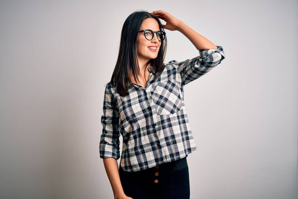 Young brunette woman with blue eyes wearing casual shirt and glasses over white background smiling confident touching hair with hand up gesture, posing attractive and fashionable - Valokuva, kuva