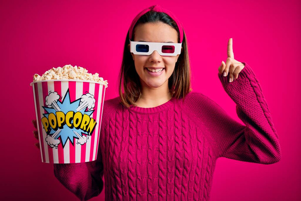 Nuori kaunis tyttö katsomassa elokuvaa käyttäen 3D-lasit syöminen laatikko popcornia hymyillen hämmästynyt ja yllättynyt ja osoittaa ylös sormilla ja nosti kädet
. - Valokuva, kuva