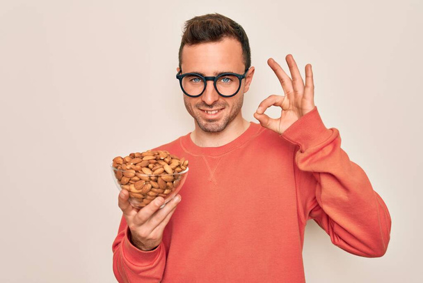 Handsome man with blue eyes holding bowl with healthy almonds snack over white background doing ok sign with fingers, excellent symbol - Valokuva, kuva