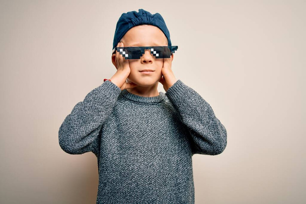 Jonge kleine blanke jongen draagt internet meme misdadiger levensbril over geïsoleerde achtergrond die lijdt aan hoofdpijn wanhopig en gestresst omdat pijn en migraine. Handen op het hoofd. - Foto, afbeelding