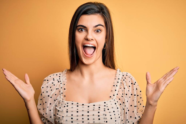 Young beautiful brunette woman wearing casual t-shirt standing over yellow background celebrating crazy and amazed for success with arms raised and open eyes screaming excited. Winner concept - Fotografie, Obrázek